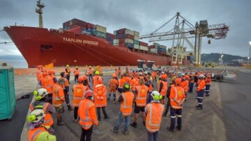 A large group of crew in Hi-Vis stand in front of a large container ship, with Cranes in the background 